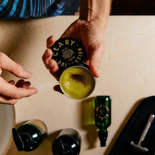 Person holding a container of Clary Sage essential oil with bottles and a tumbler on a table.