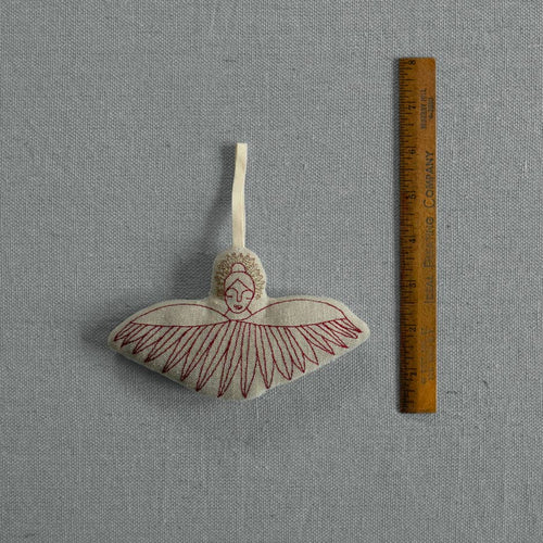 A small embroidered angel ornament with outstretched wings and a hanging loop, shown next to a wooden ruler on a gray fabric background.