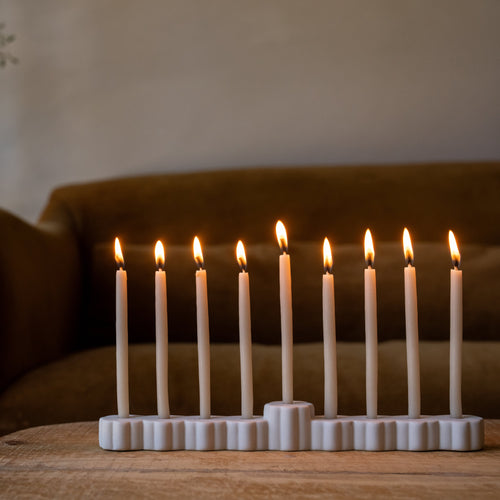 A nine-light Hanukkah menorah made of matte white ceramic, holding nine lit white taper candles, set on a wooden table in front of a brown leather sofa.