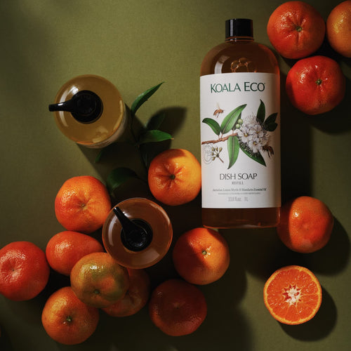 A bottle of KOALA ECO DISH SOAP stands upright on a dark olive green surface, surrounded by oranges and a sliced orange, with two amber drink cups with black straws nearby. The product label shows a white background with green leaves and white blossoms.