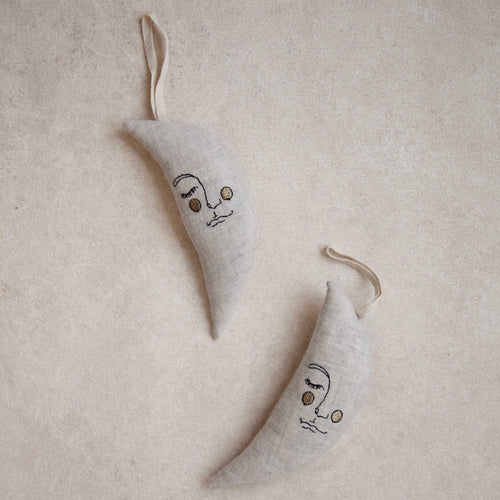Two small crescent moon-shaped fabric hanging ornaments in a light beige linen-like material, each embroidered with a simple face and shown on a textured beige background.