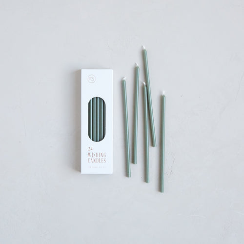 24 wishing candles in moss green photographed next to white packaging box. Skinny tall taper candles can be used as birthday candles or meditation candles.