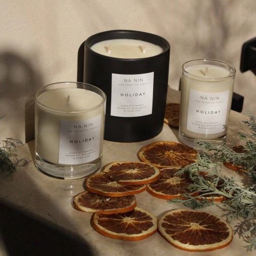 Three NA'NIN candles with 'Holiday' labels are styled on a marble surface with dried orange slices and greenery under warm, dramatic sunlight and shadows.