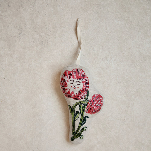 A small handmade felt ornament shaped like a flower with a smiling face, red and pink petals, and green stem details, hanging against a textured beige background.