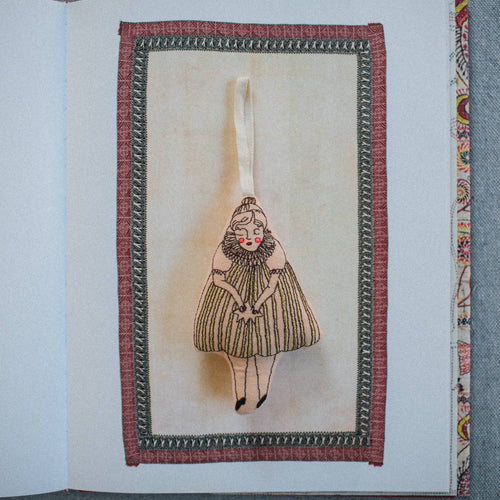 A handmade fabric bookmark or ornament featuring an embroidered illustration of a woman in a pleated dress, suspended by a white ribbon. The design is framed by a decorative border with a red outer edge and a dark patterned inner edge, set against an open book page.