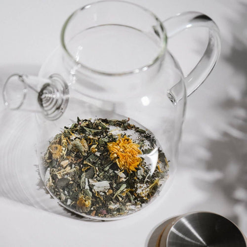 Clear glass teapot with herbs and flowers on a white surface