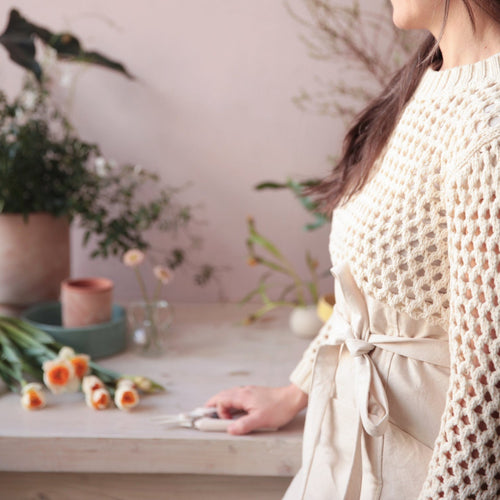 Person wearing a beige knit sweater and apron, standing in a room with flowers and plants.